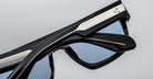 Close-up of two Jacques Marie Mage Ritz sunglasses with blue-tinted lenses, stacked on a light gray background. The black frames have metallic accents on the temples.