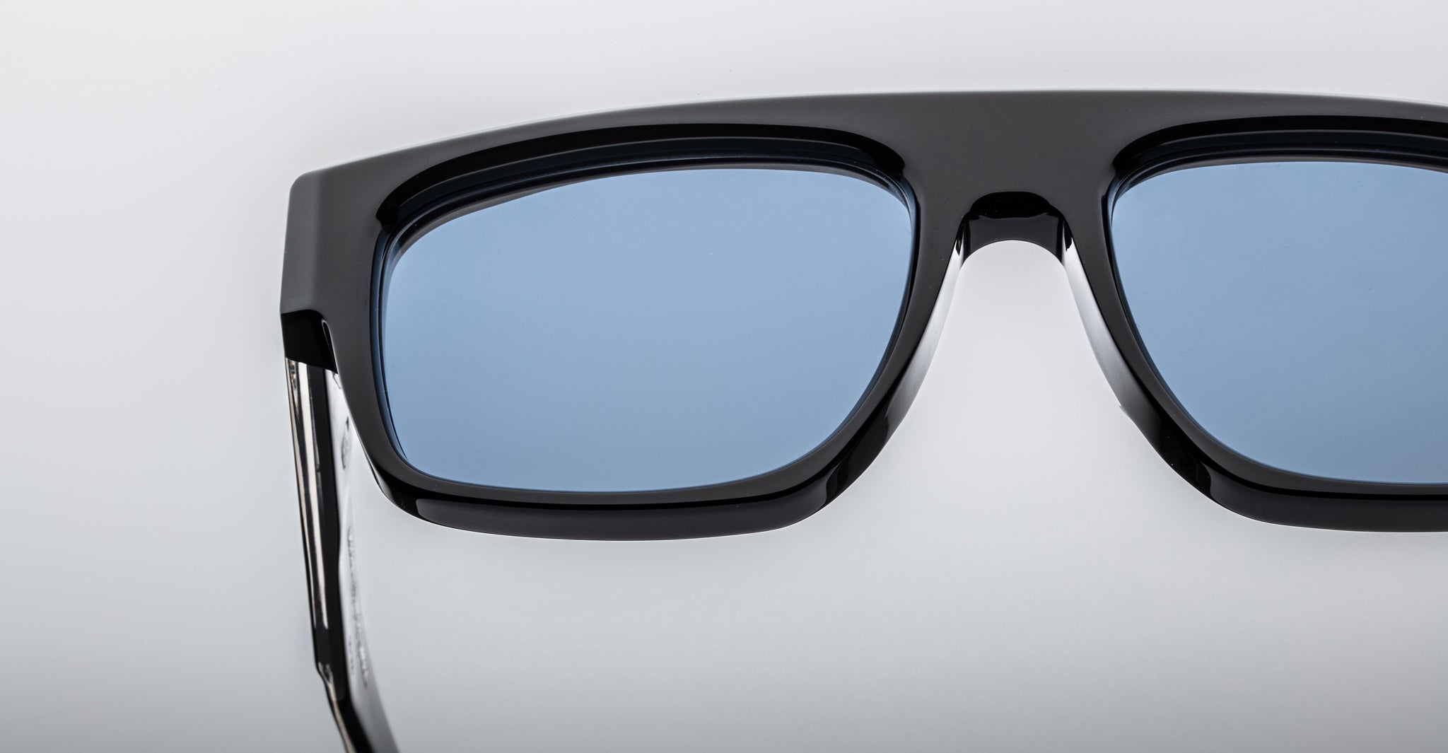 Close-up of the upper half of Jacques Marie Mage Ritz sunglasses in black with blue-tinted lenses, set against a plain white background. Only part of the frame and one arm are visible.