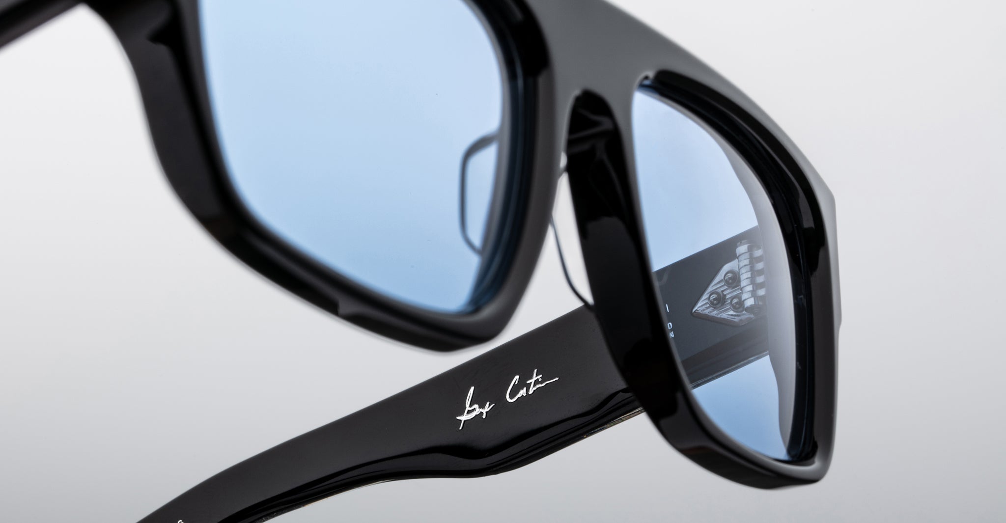 Close-up of Jacques Marie Mage Ritz black sunglasses with blue-tinted lenses, featuring a handwritten-style signature on the arm and a triangular logo seen through the lens against a plain light background.