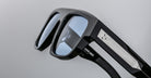 A close-up of Jacques Marie Mage Ritz black eyeglass frames with light blue-tinted lenses and a silver ribbed metal temple accent featuring a small engraved symbol.