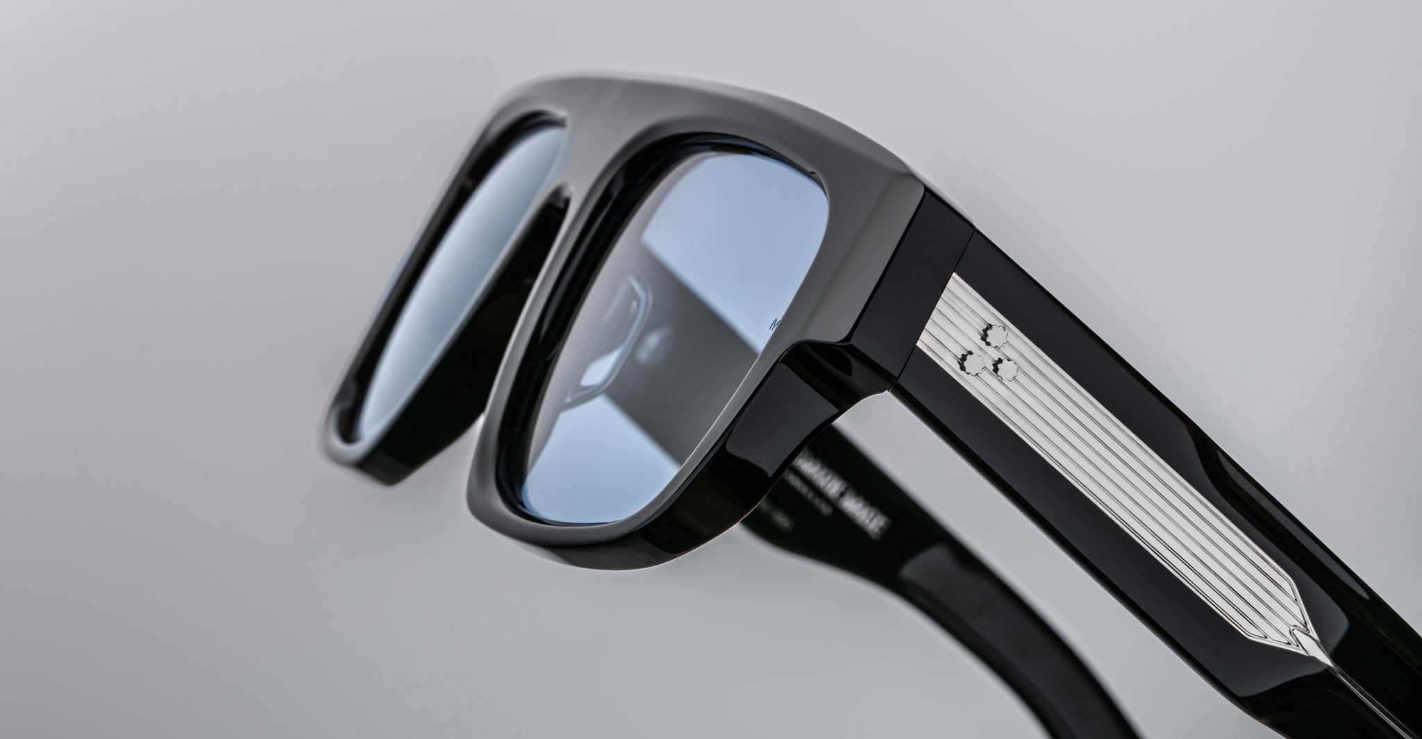 A close-up of Jacques Marie Mage Ritz black eyeglass frames with light blue-tinted lenses and a silver ribbed metal temple accent featuring a small engraved symbol.
