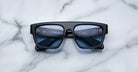 A pair of Jacques Marie Mage Ritz black rectangular sunglasses with thick frames and dark lenses rests on a white marble surface, with reflections visible on the lenses.