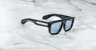 Jacques Marie Mage Ritz rectangular black sunglasses with blue-tinted lenses rest on white marble. The frame features a metallic accent and small cross on the temple, with a signature visible inside one arm.