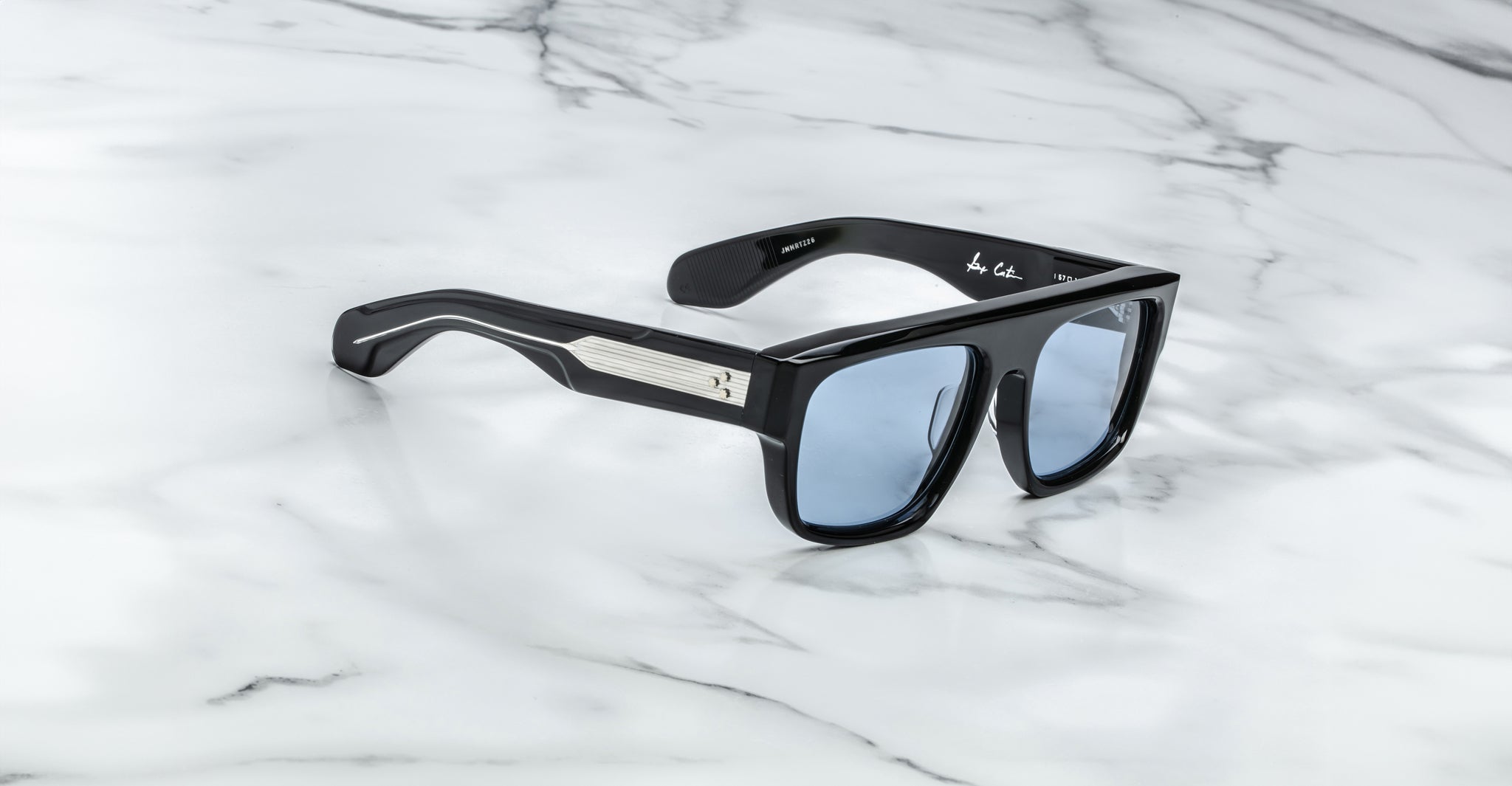 Jacques Marie Mage Ritz rectangular black sunglasses with blue-tinted lenses rest on white marble. The frame features a metallic accent and small cross on the temple, with a signature visible inside one arm.