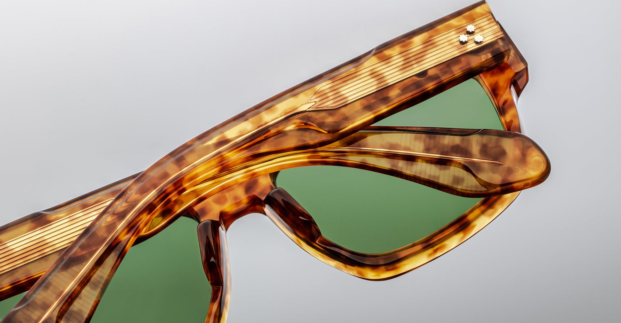 Close-up of Jacques Marie Mage Ritz sunglasses featuring a tortoiseshell frame, green lenses, and two small metallic dots on the upper corners, set against a plain light gray background.