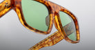 Close-up of Jacques Marie Mage Ritz sunglasses featuring a tortoiseshell frame, green lenses, and a white signature on the temple, set against a neutral light gray background.