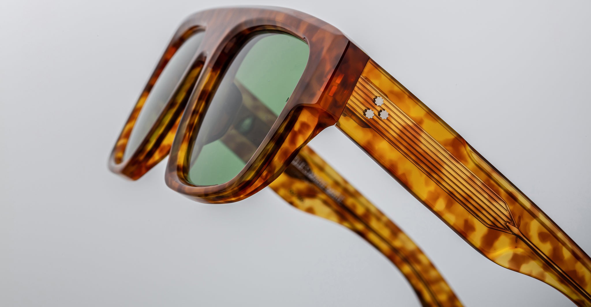 Close-up of Jacques Marie Mage Ritz sunglasses featuring a tortoiseshell frame, green lenses, gold detailing, and two small white accents on the temple against a neutral background.