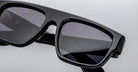 Close-up of Jacques Marie Mage Ritz, modern black rectangular sunglasses with dark tinted lenses on a white background. Subtle branding is visible on the inside of the frame.