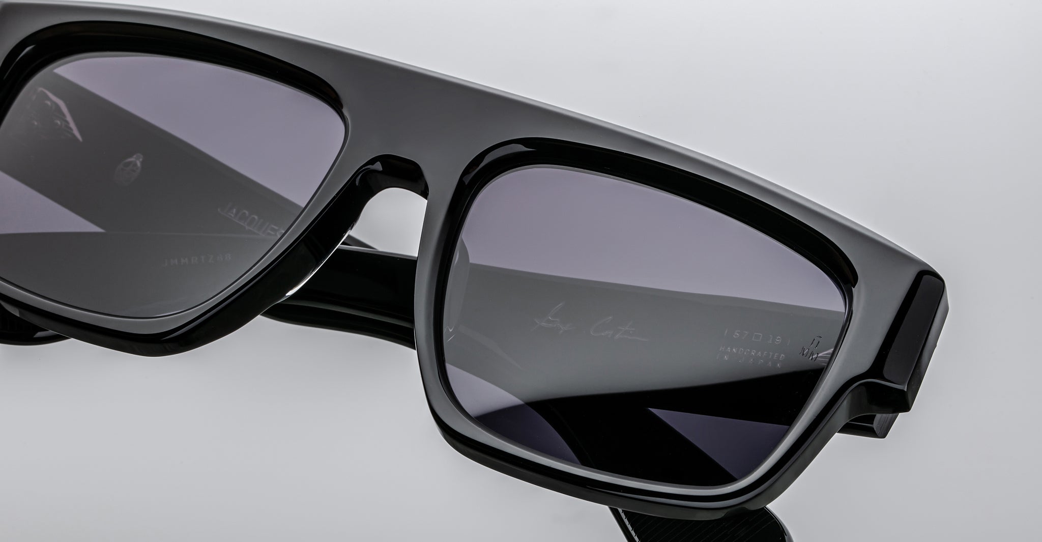 Close-up of Jacques Marie Mage Ritz, modern black rectangular sunglasses with dark tinted lenses on a white background. Subtle branding is visible on the inside of the frame.
