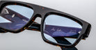 Close-up of Jacques Marie Mage Ritz tortoiseshell eyeglasses with blue-tinted lenses. Reflections reveal the interior frames, displaying white text with model numbers, brand name, and “handcrafted in Japan.”.