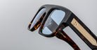 Close-up of Jacques Marie Mage Ritz eyeglasses featuring thick brown tortoiseshell frames, blue-tinted lenses, and gold detailing on the arms, set against a plain light gray background.