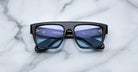 The Jacques Marie Mage Ritz sunglasses, featuring a black rectangular frame and blue-tinted lenses, rest face-up on a white marble surface with their inner arms and brand details clearly visible.
