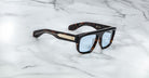 The Jacques Marie Mage Ritz are thick tortoiseshell sunglasses with blue-tinted lenses and gold accents on the temples, displayed on a white marble surface.