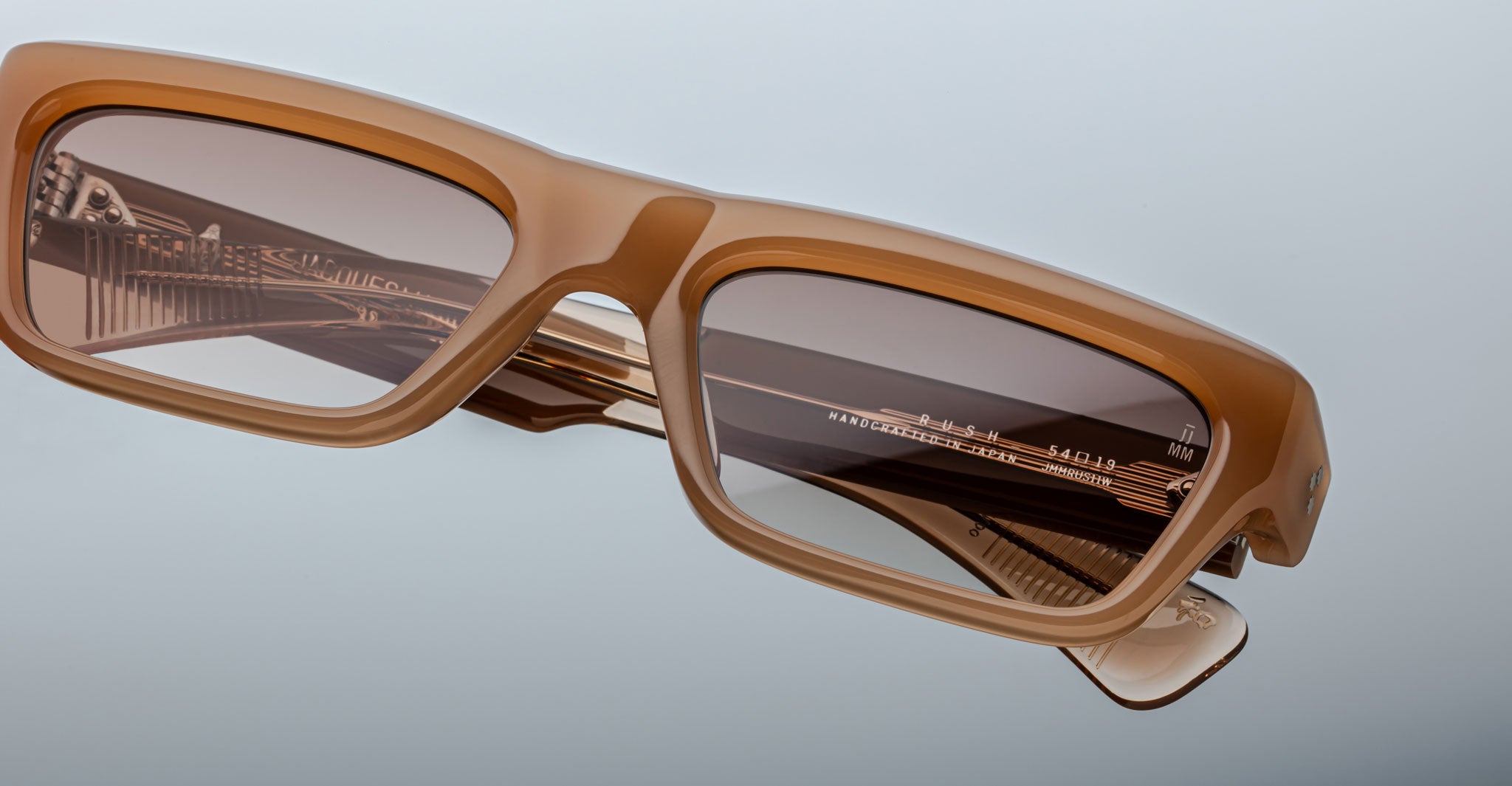 A close-up of Jacques Marie Mage Rush brown rectangular sunglasses with thick frames and tinted lenses, angled on a light gray background. Gold detailing and text appear on the inside arm.