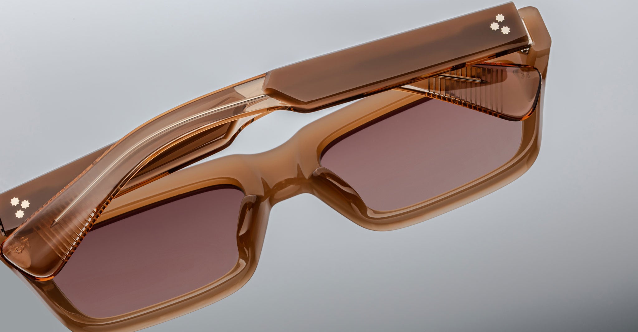 The Jacques Marie Mage Rush sunglasses, featuring thick brown rectangular frames with tinted lenses and small white flower designs on the hinges, rest on a reflective grey surface.