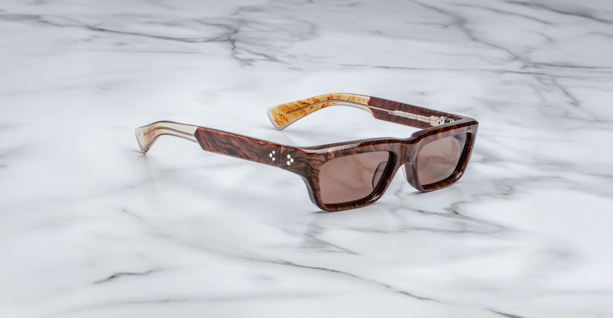 The Jacques Marie Mage Rush sunglasses, featuring thick, brown marbled rectangular frames and brown-tinted lenses, rest on a white marble surface.