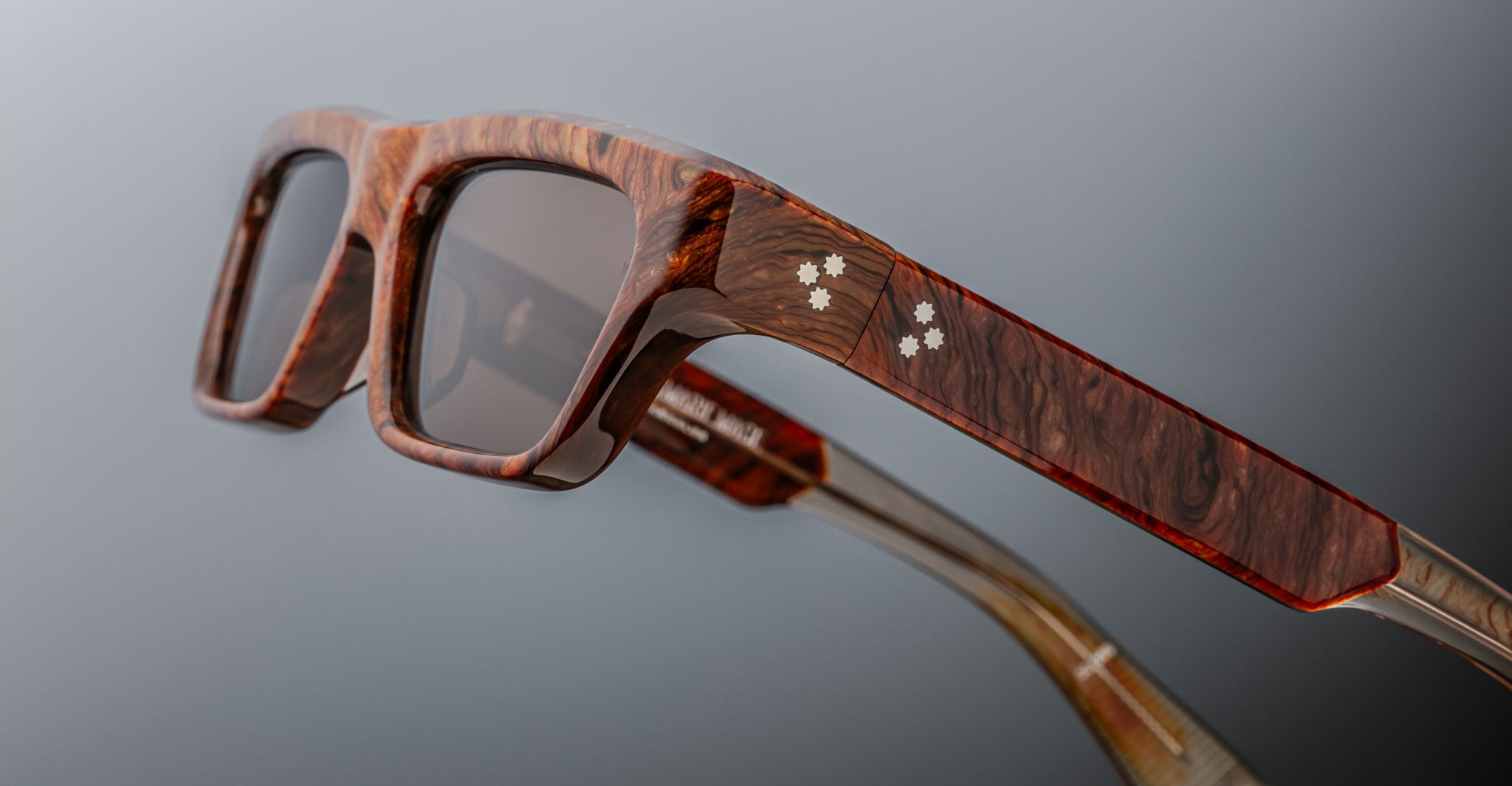 Close-up of Jacques Marie Mage Rush sunglasses featuring wooden frames, rectangular lenses, and star-shaped inlays on the temples, set against a neutral gradient background.