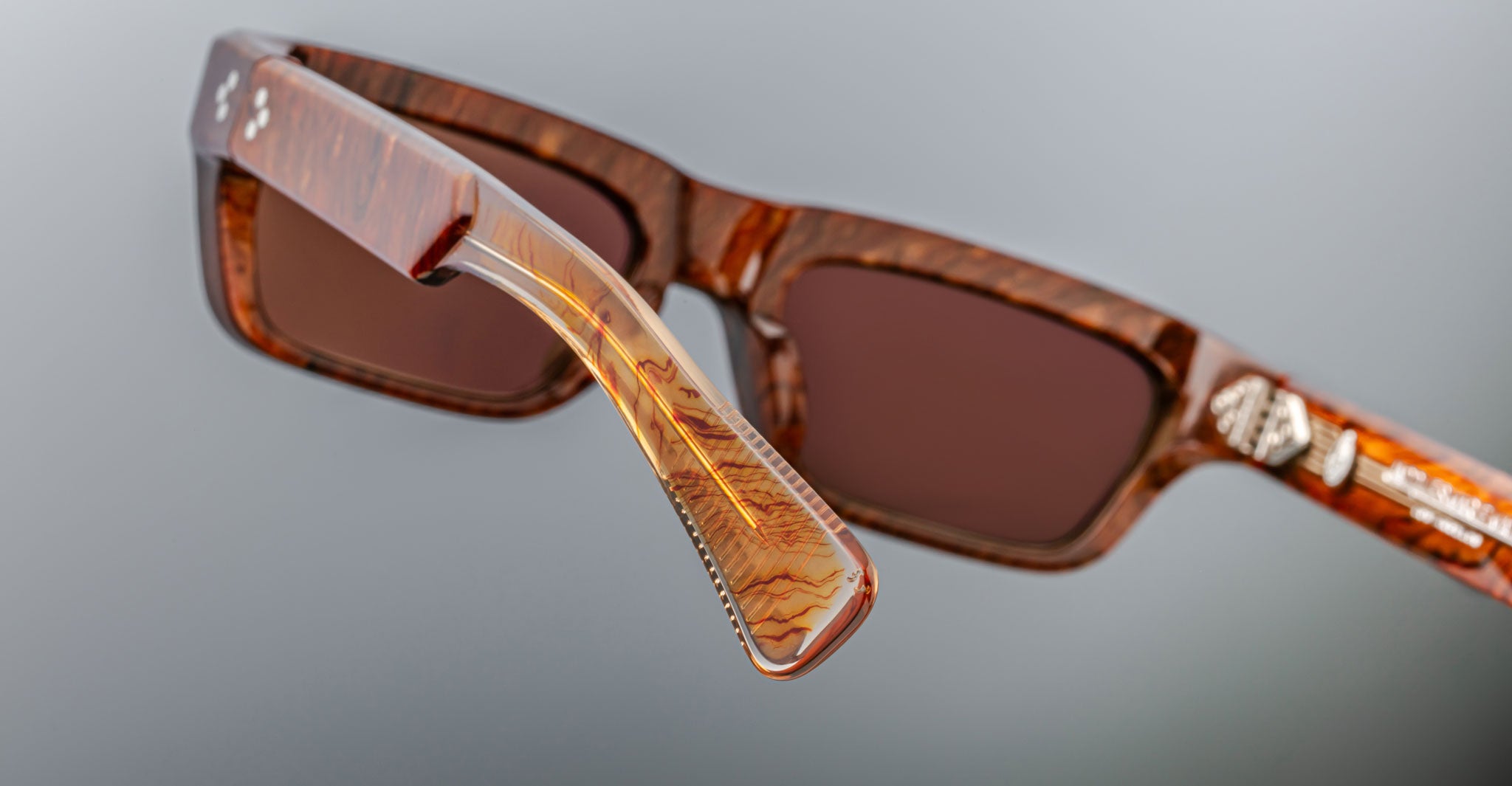 Close-up of Jacques Marie Mage Rush brown rectangular sunglasses with marbled frames and arms. The image highlights the hinge and detailed texture of the arm against a neutral gray background.