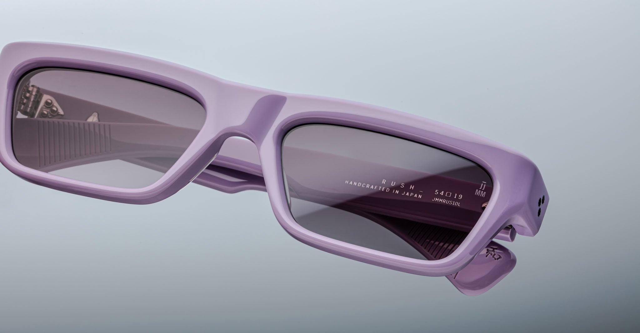 Close-up of Jacques Marie Mage Rush sunglasses in pastel purple with rectangular frames and dark tinted lenses. Product and manufacturing details appear inside the right temple arm, set against a plain light gradient background.