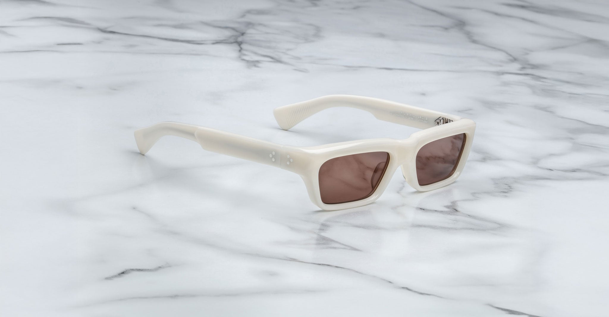 The Jacques Marie Mage Rush sunglasses, featuring thick off-white rectangular frames and brown-tinted lenses, rest on a white marble surface.
