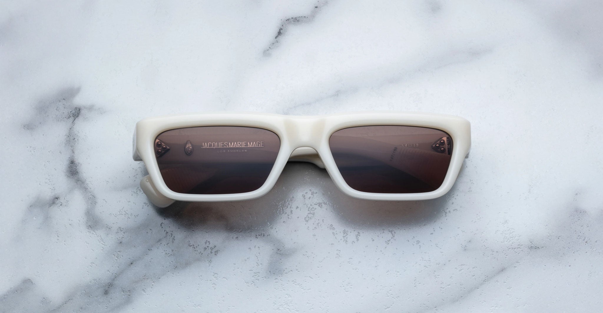 The Jacques Marie Mage Rush sunglasses, featuring thick white rectangular frames and dark lenses, are displayed on a white marble surface.