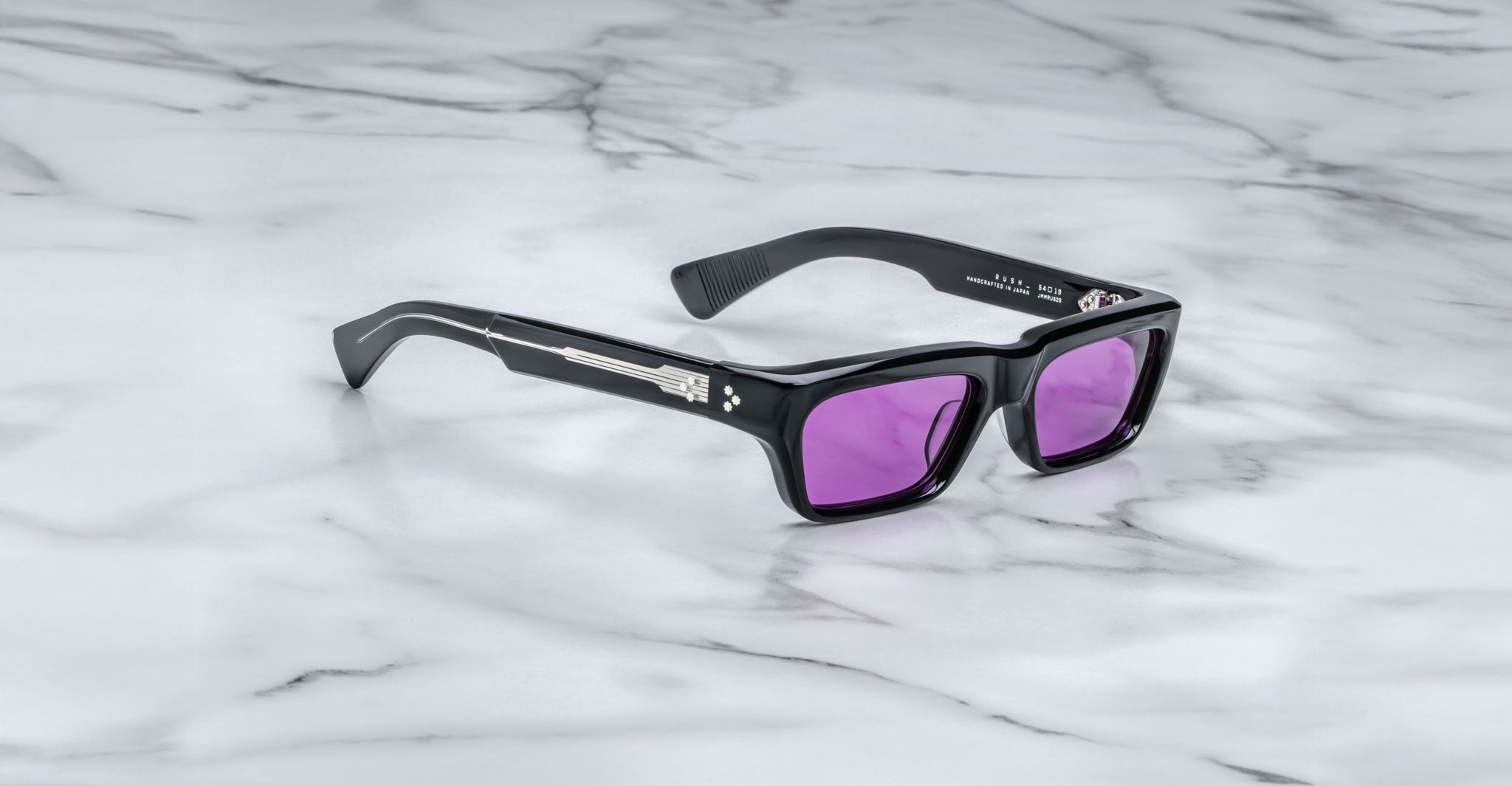 The Jacques Marie Mage Rush sunglasses, featuring a black rectangular frame and purple-tinted lenses with silver detailing on the arms, rest on a white marble surface.