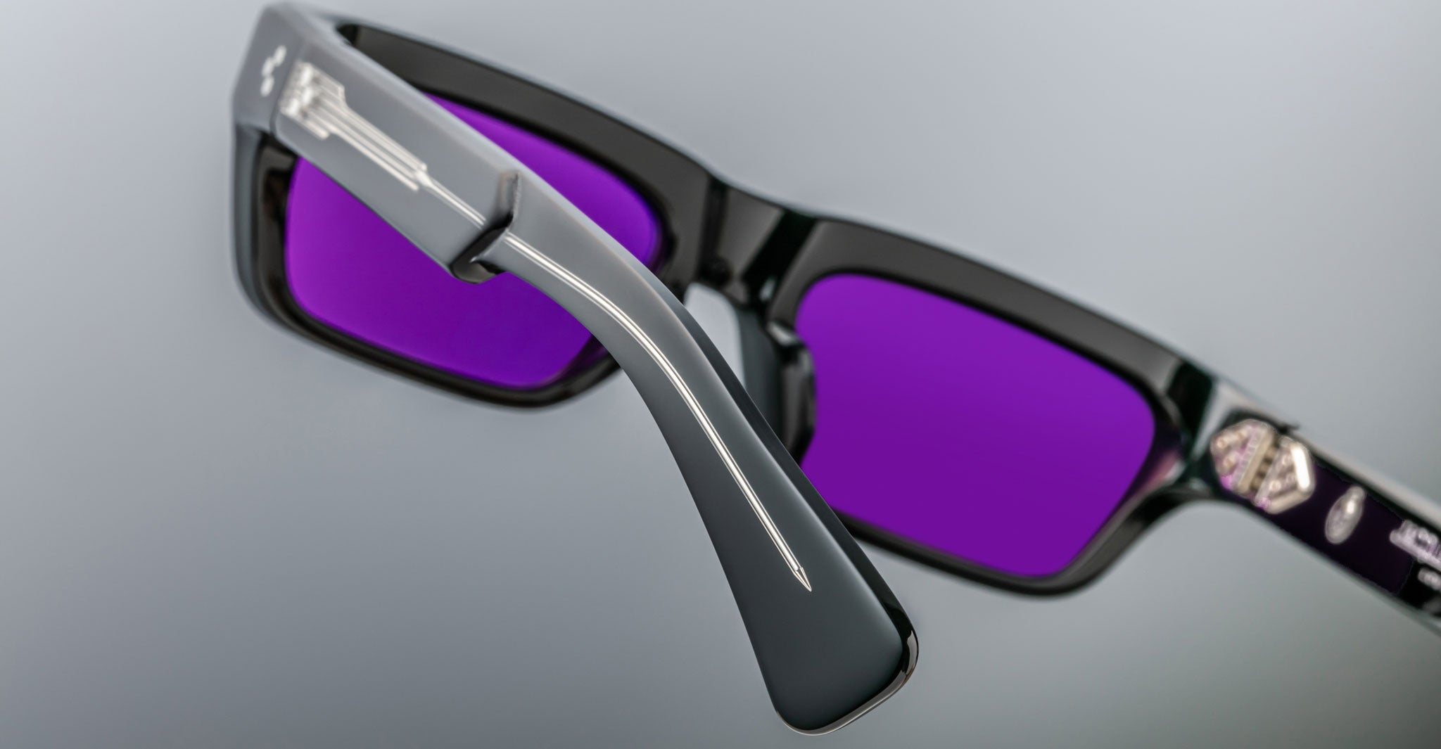 Close-up of Jacques Marie Mage Rush black rectangular sunglasses with purple-tinted lenses; focus highlights the detailed texture of the temple arms and lens against a neutral background.