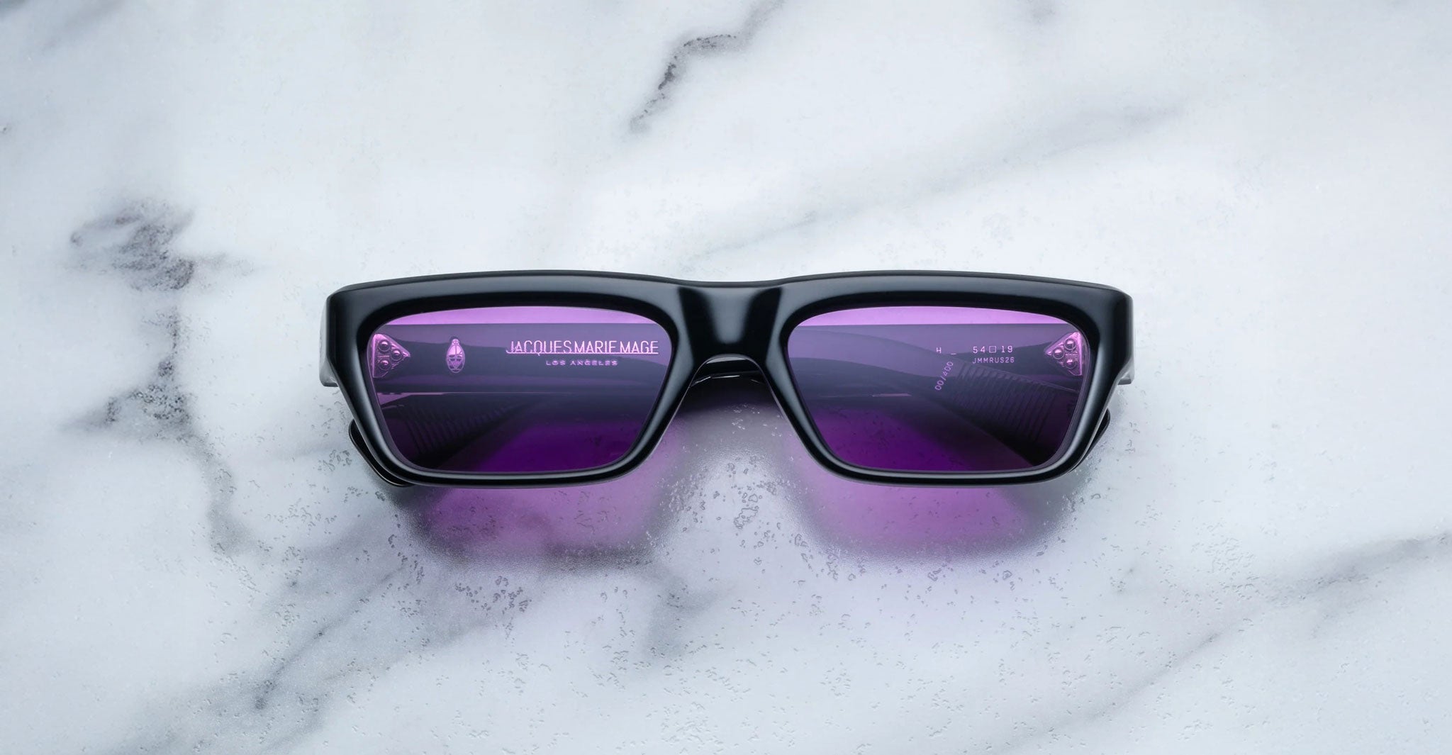 A pair of Jacques Marie Mage Rush black rectangular sunglasses with purple-tinted lenses rests on a white marble surface. Reflections in the lenses hint at partial text and shapes, suggesting the photo was taken indoors.