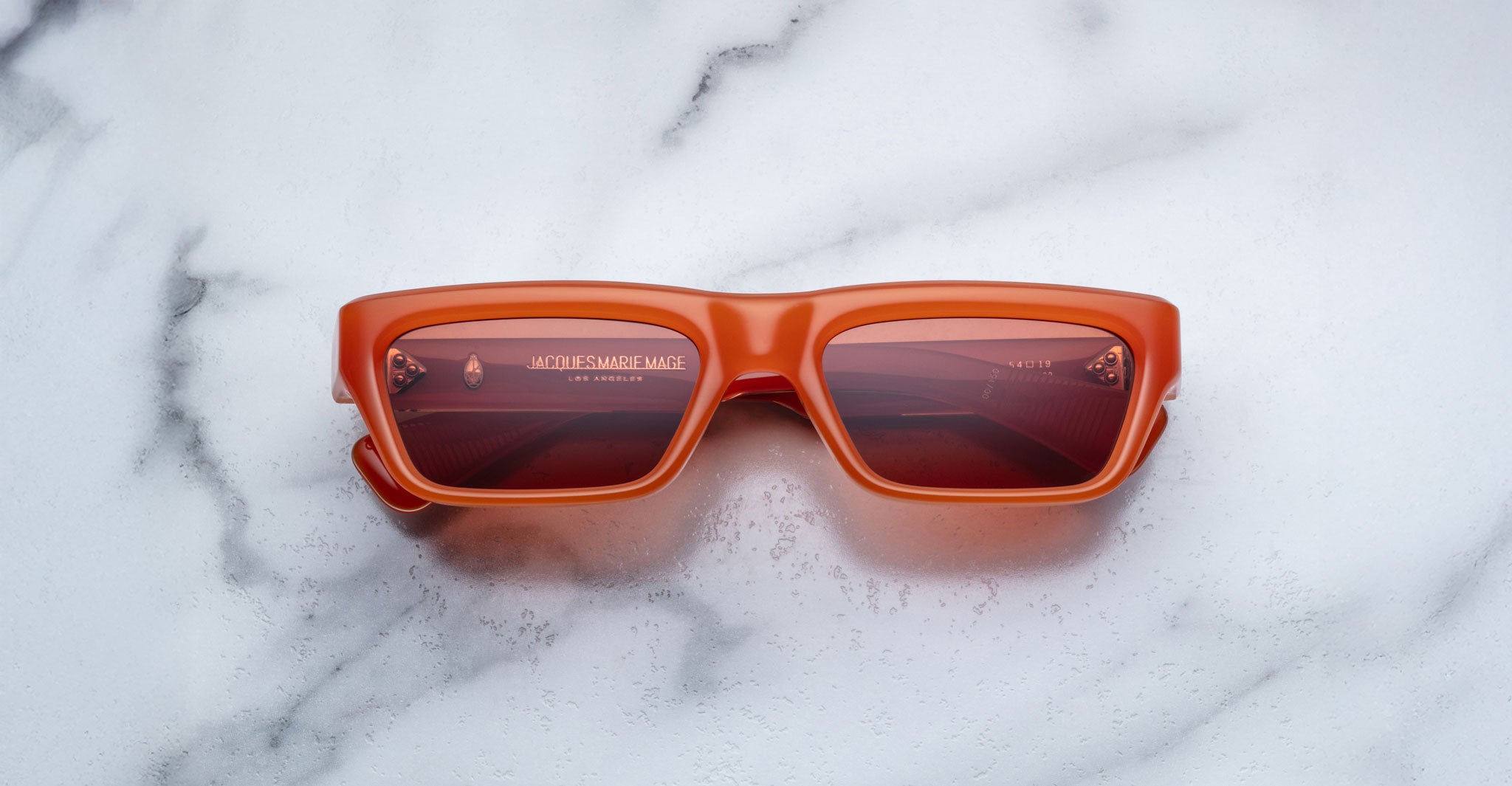 Jacques Marie Mage Rush rectangular sunglasses with thick orange frames and red-tinted lenses rest on a white marble surface. JACQUES MARIE MAGE and LOS ANGELES are visible on the inside of the left lens.