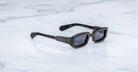Jacques Marie Mage Ryder in Black, rectangular black acetate sunglasses shown from a side angle, featuring thick sculpted rims, wide temples with stepped detailing, and dark tinted lenses on a light marble surface.