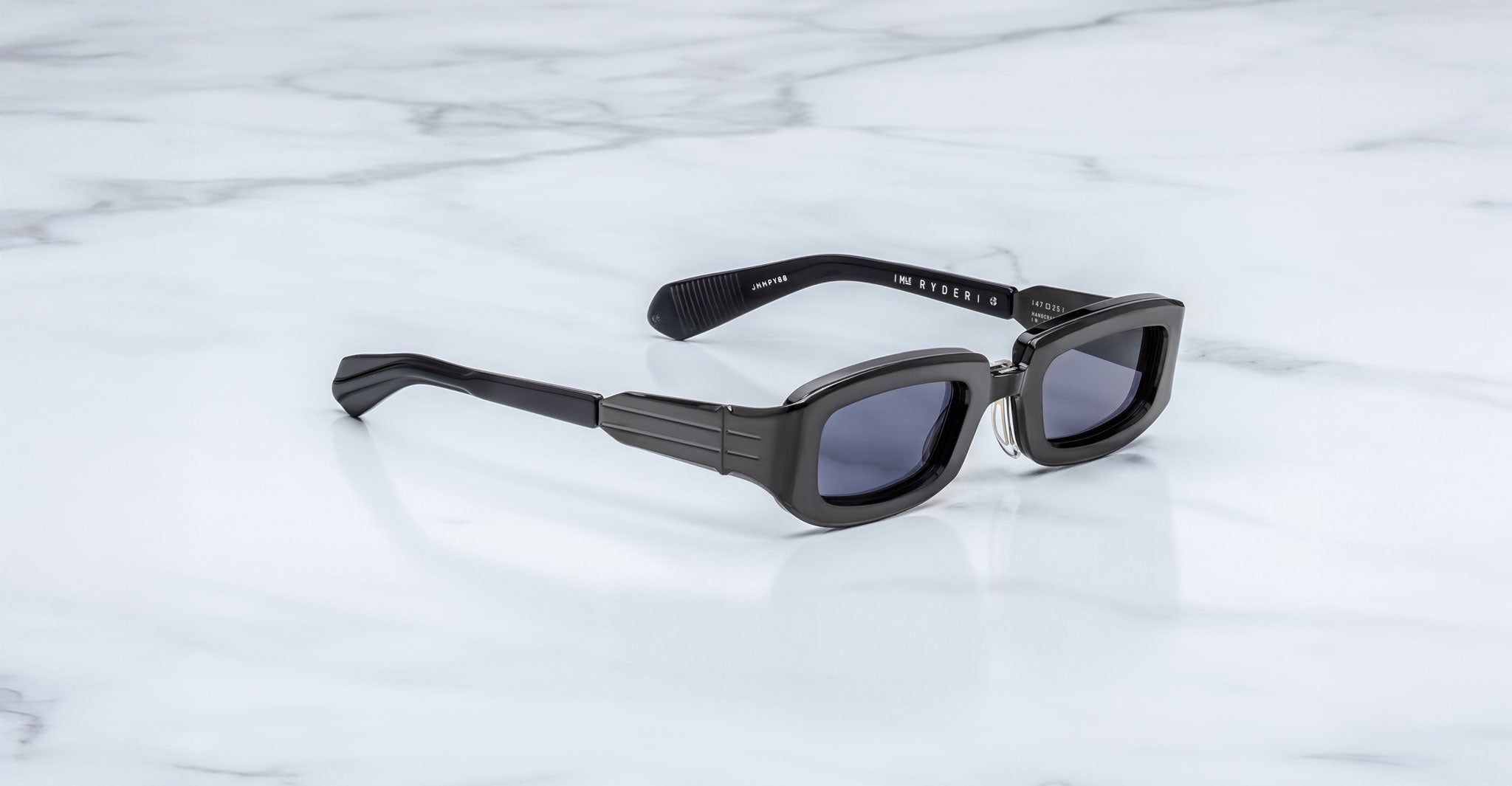 Jacques Marie Mage Ryder in Black, rectangular black acetate sunglasses shown from a side angle, featuring thick sculpted rims, wide temples with stepped detailing, and dark tinted lenses on a light marble surface.