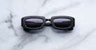 Jacques Marie Mage Ryder in Black, rectangular black acetate sunglasses with thick sculpted rims, a low-profile bridge, and dark tinted lenses, shown front-facing on a light marble surface.