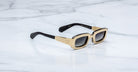 Jacques Marie Mage Ryder in Gold, rectangular gold-tone acetate sunglasses shown from a side angle, featuring thick sculpted rims, wide temples with stepped detailing, and gradient tinted lenses on a light marble surface.
