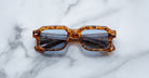 A pair of Jacques Marie Mage Sandro square tortoiseshell sunglasses with blue-tinted lenses rests on a white marble surface.