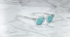 Jacques Marie Mage Sandro clear-framed sunglasses with teal lenses placed on a white marble surface featuring gray veining.