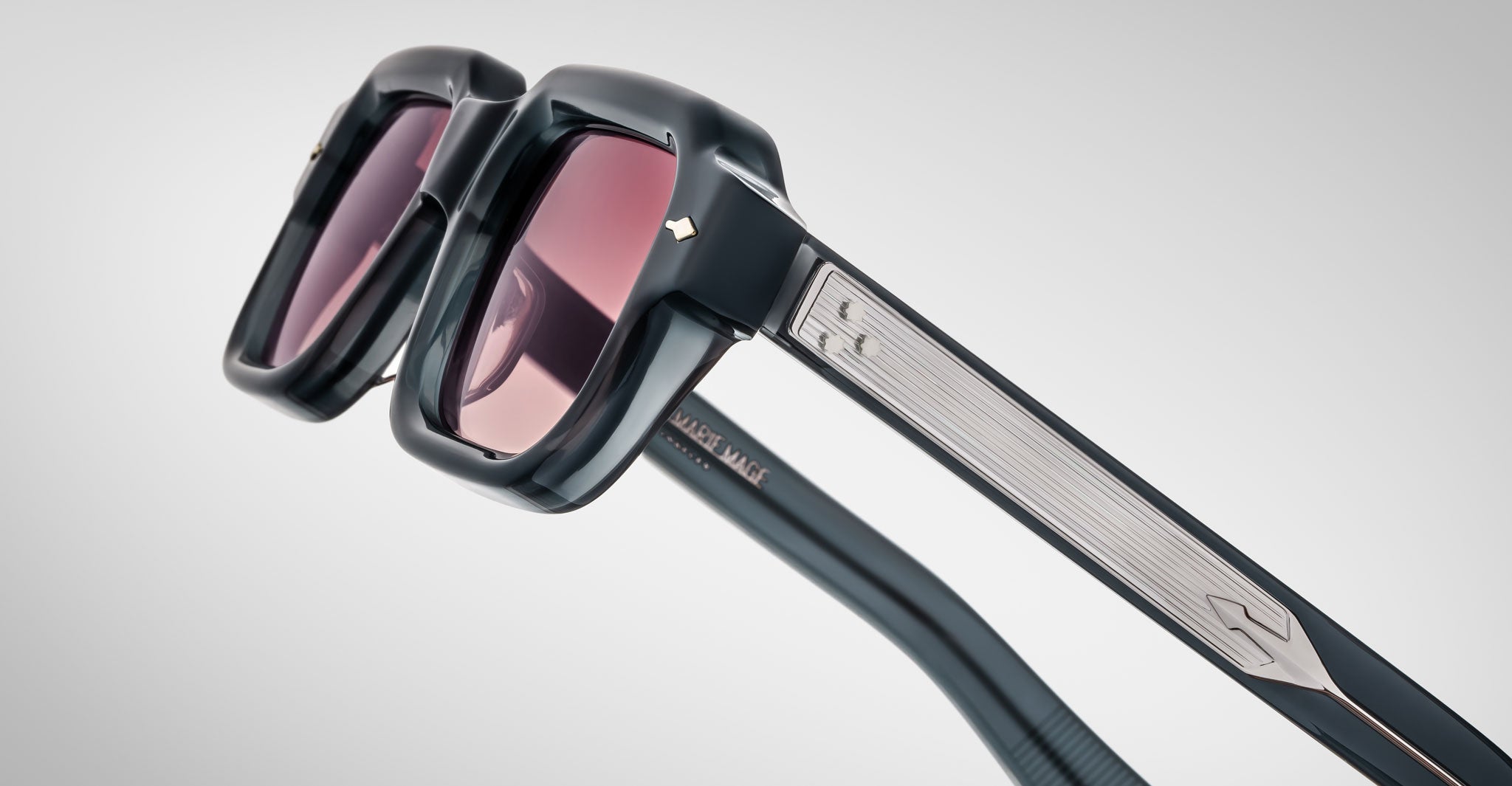 Close-up of Jacques Marie Mage Sandro sunglasses: black, thick frames with pink-tinted lenses. The arms display metallic accents, a textured pattern, and a small metallic logo near the hinge.