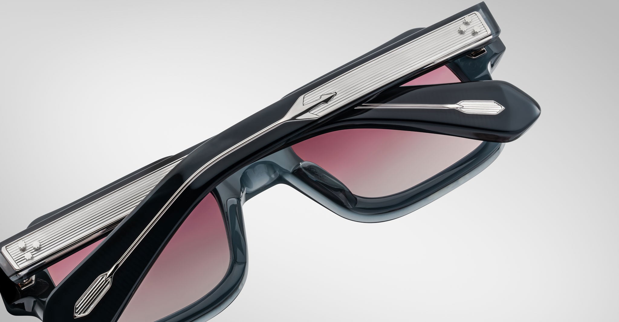 A close-up of Jacques Marie Mage Sandro sunglasses with thick, dark frames and pink-tinted lenses. The metal arms have a textured pattern and arrow-shaped detail near the hinges, set against a plain light gray background.