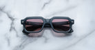 A pair of Jacques Marie Mage Sandro square sunglasses with black frames and dark red lenses rests on a white marble surface with grey veining.