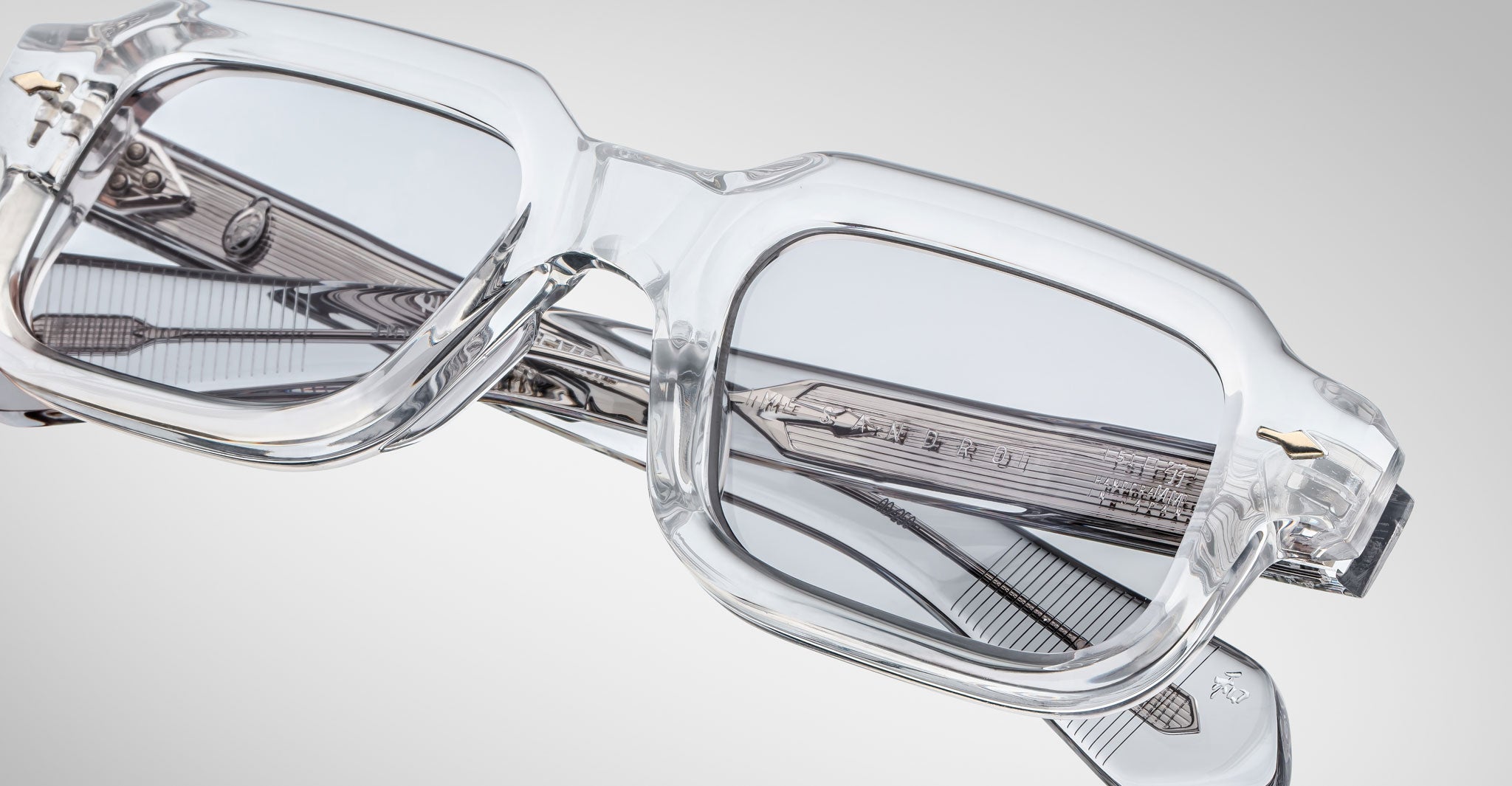Close-up of Jacques Marie Mage Sandro clear rectangular eyeglass frames with gray-tinted lenses and intricate arm detailing, set against a light gray background.