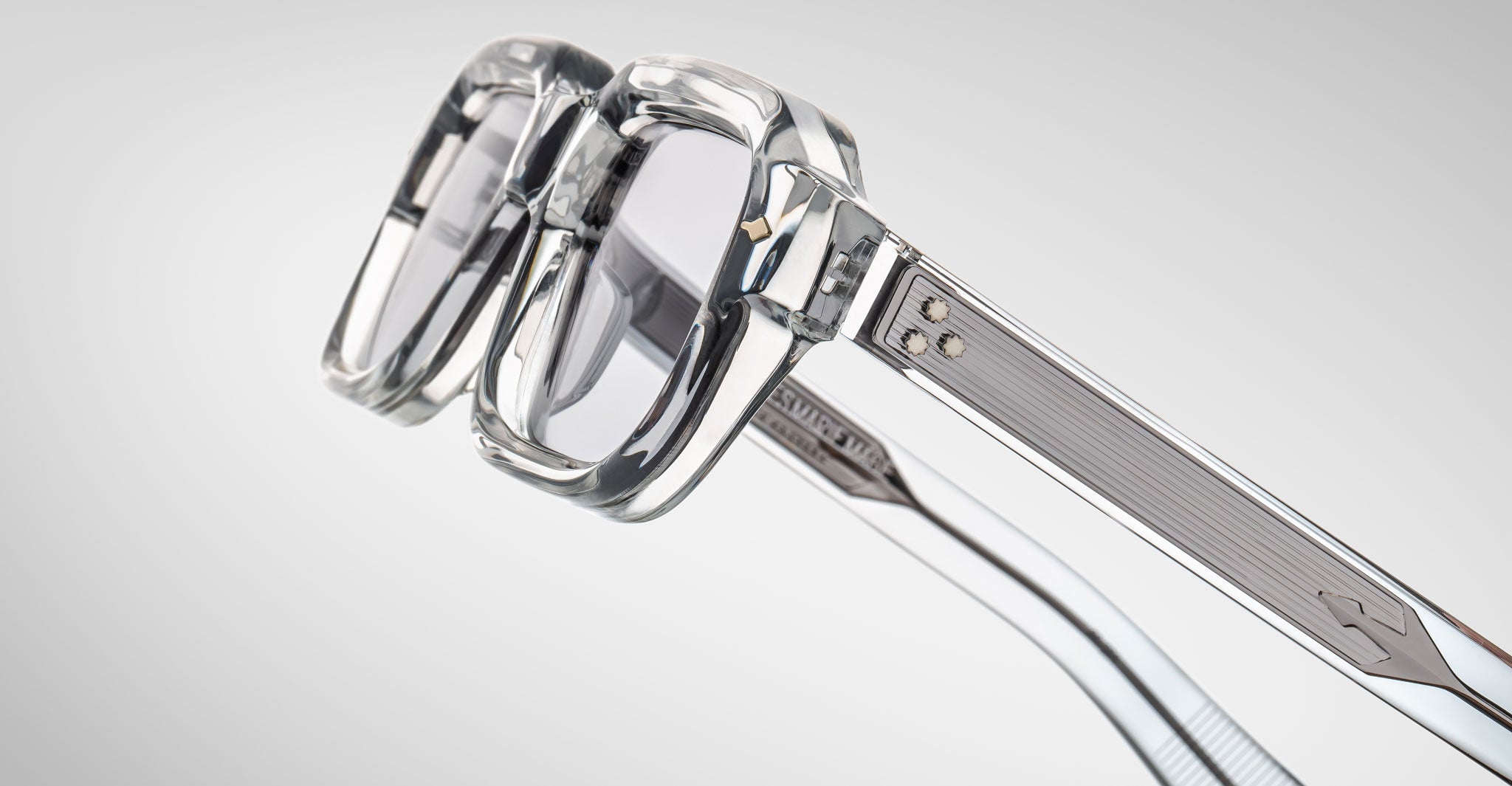 Close-up view of Jacques Marie Mage Sandro eyeglasses featuring clear, thick rims, transparent lenses, and metallic accents on the arms, set against a light gray background.