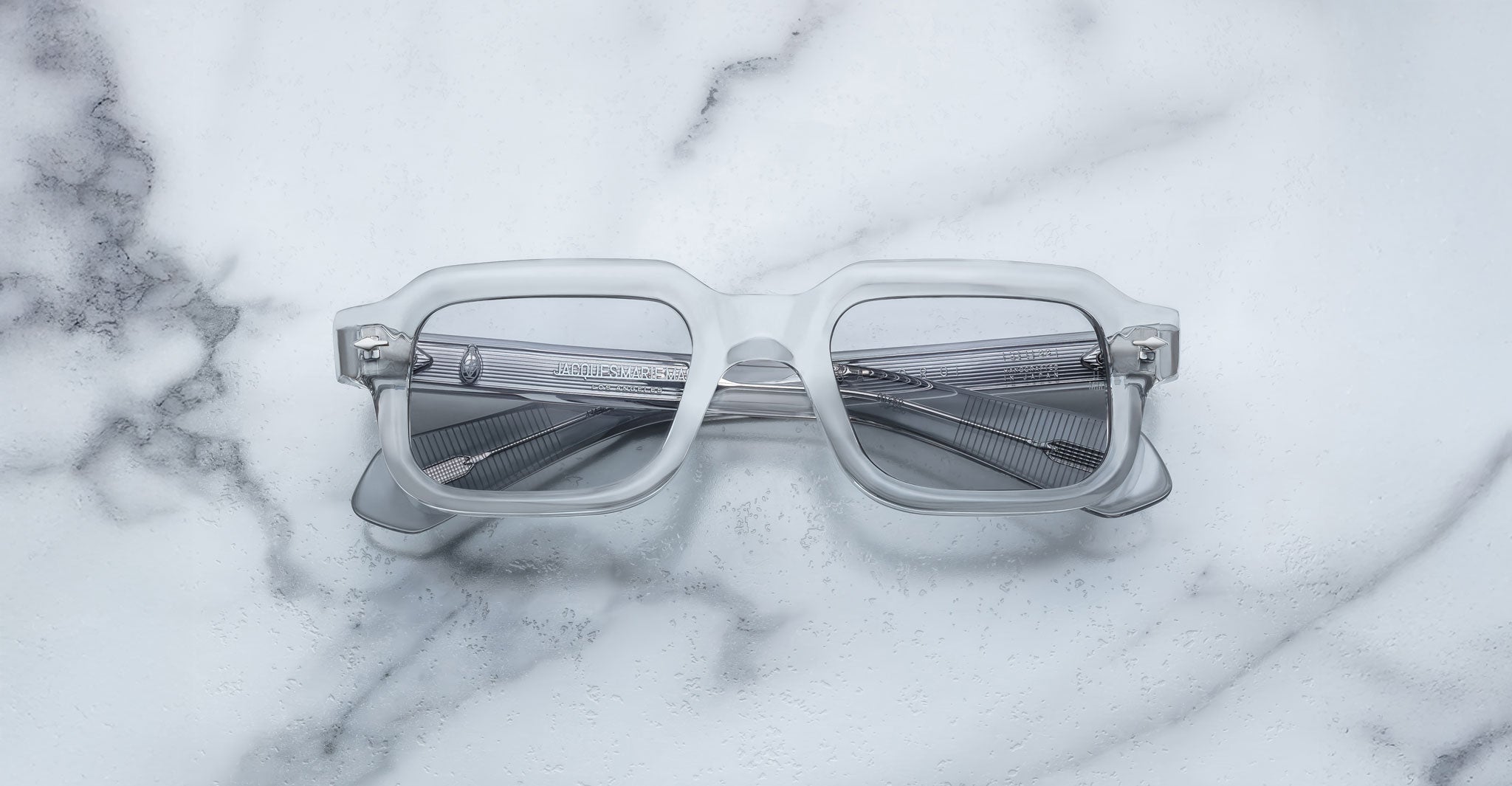 The Jacques Marie Mage Sandro eyeglasses, featuring clear rectangular frames and grey-tinted lenses, rest on a white marble surface with grey veining.