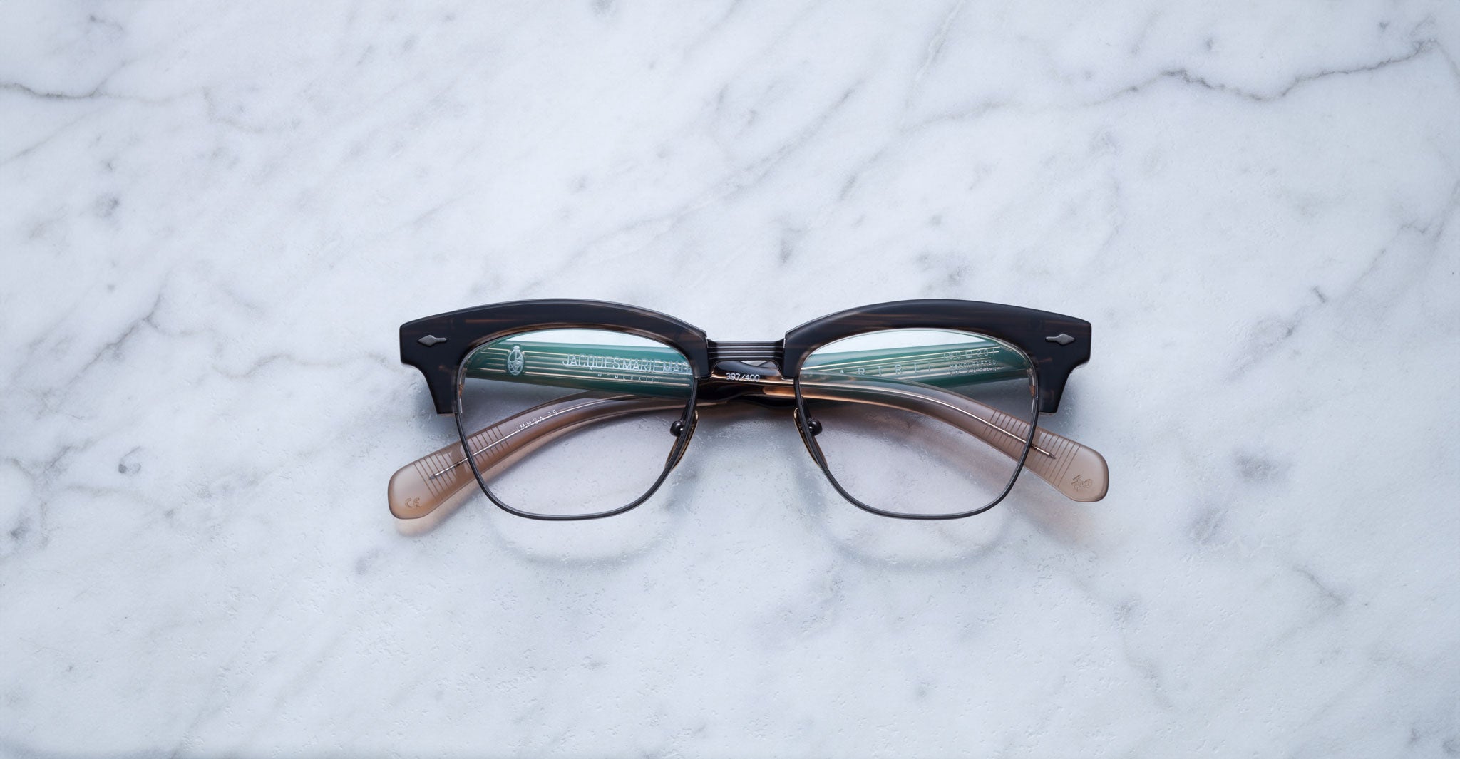 A pair of Jacques Marie Mage Sartre glasses with black and translucent brown clubmaster optical frames rests on a white marble surface.