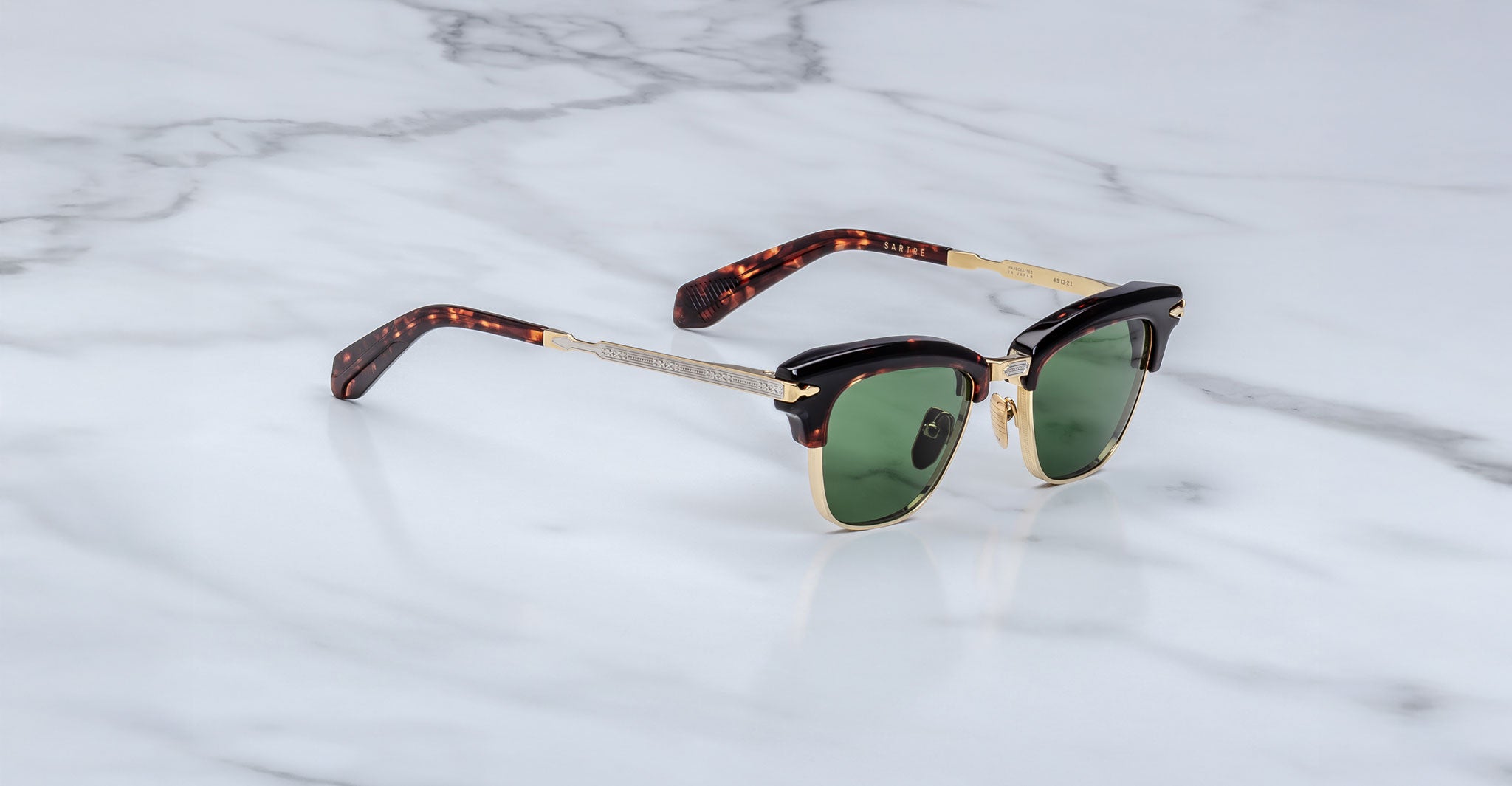 A pair of Jacques Marie Mage Sartre glasses with gold metal frames, tortoiseshell-patterned arms, and green lenses sits on a white marble surface.
