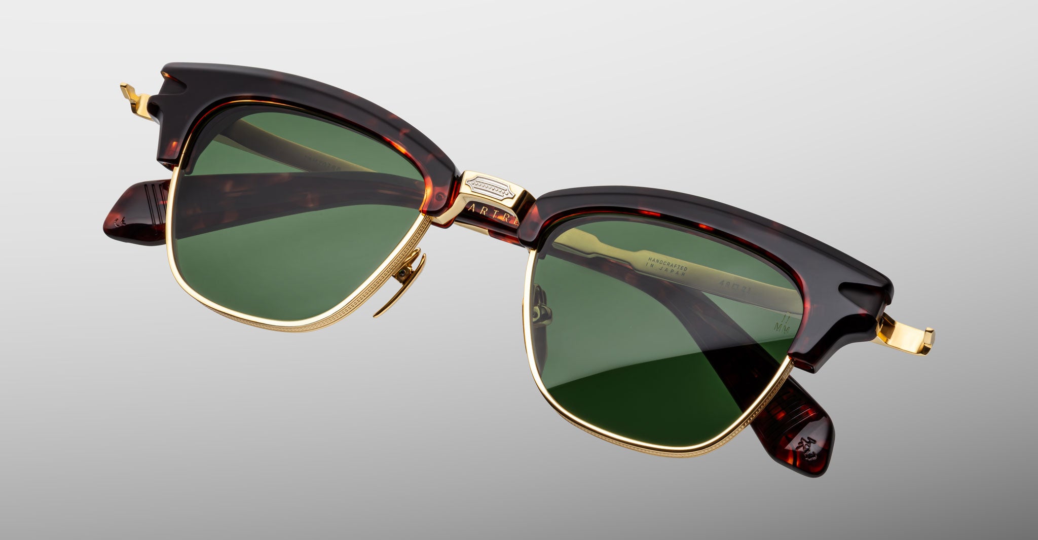 The Jacques Marie Mage Sartre sunglasses by Jacques Marie Mage feature tortoiseshell and gold frames with green lenses, inspired by classic clubmaster design, displayed on a neutral gray background.