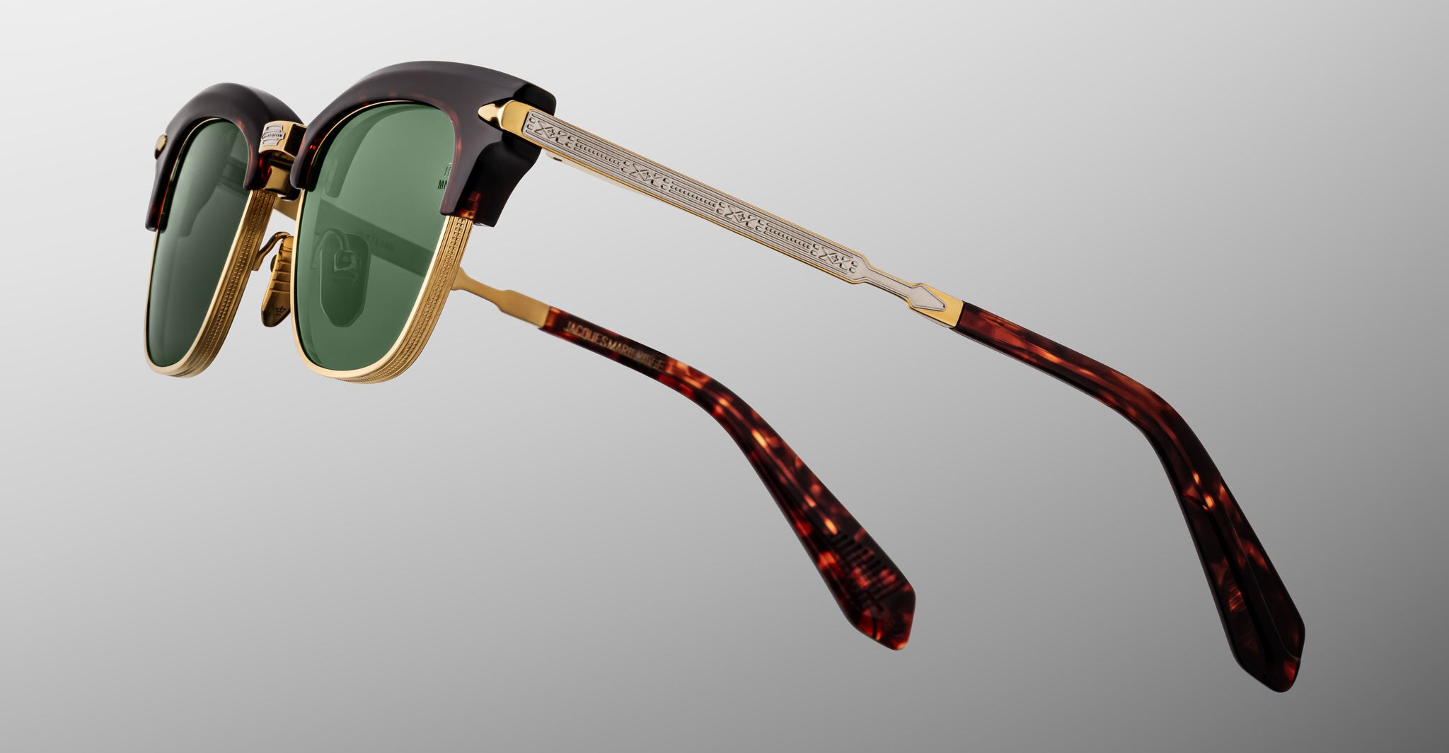 Jacques Marie Mage Sartre glasses with green lenses, gold metal accents, engraved silver arms, and tortoiseshell-patterned temple tips are shown on a gray background.