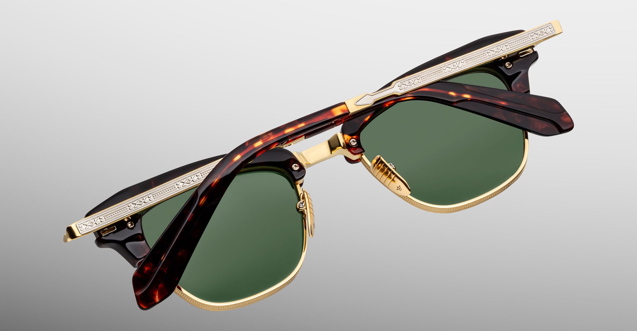 A pair of Jacques Marie Mage Sartre glasses with gold frames, green lenses, and tortoiseshell-patterned arms is displayed folded against a light gray background.