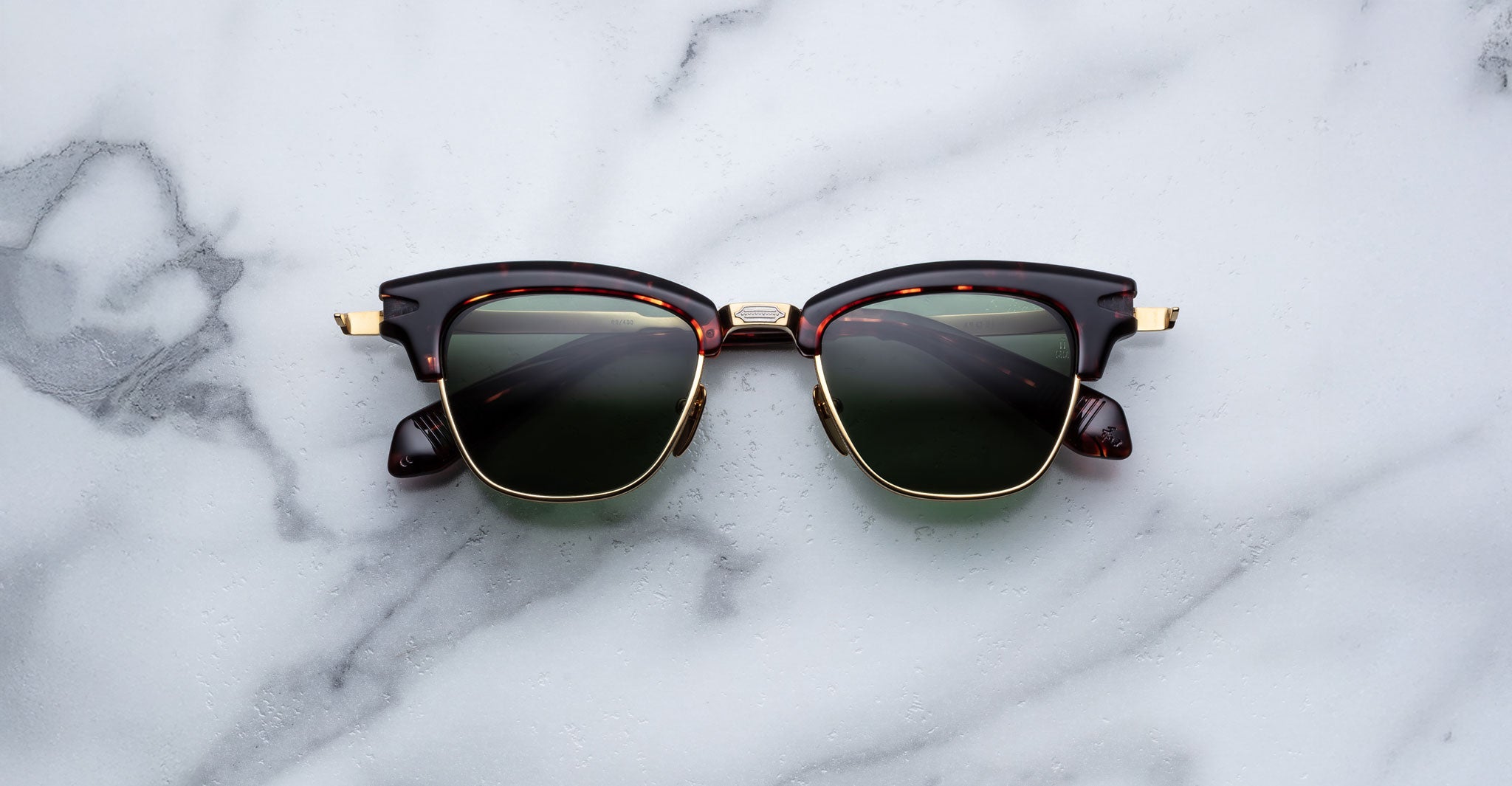 A pair of Jacques Marie Mage Sartre glasses in brown and gold with dark lenses rests on a white marble surface.
