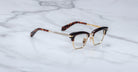 The Jacques Marie Mage Sartre glasses by Jacques Marie Mage feature gold rims, black upper frames, and tortoiseshell-patterned arms in a classic clubmaster style, shown on a white marble surface.