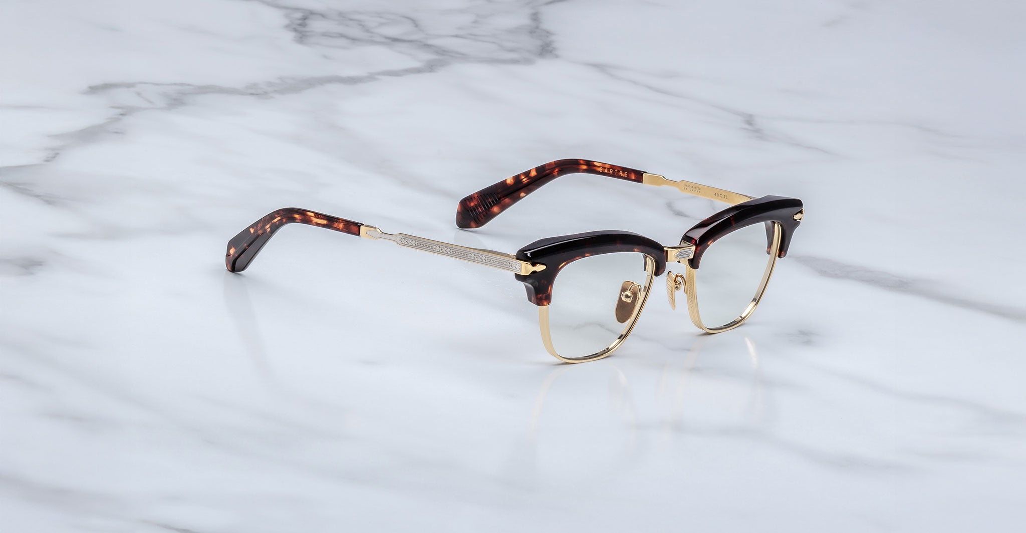 The Jacques Marie Mage Sartre glasses by Jacques Marie Mage feature gold rims, black upper frames, and tortoiseshell-patterned arms in a classic clubmaster style, shown on a white marble surface.