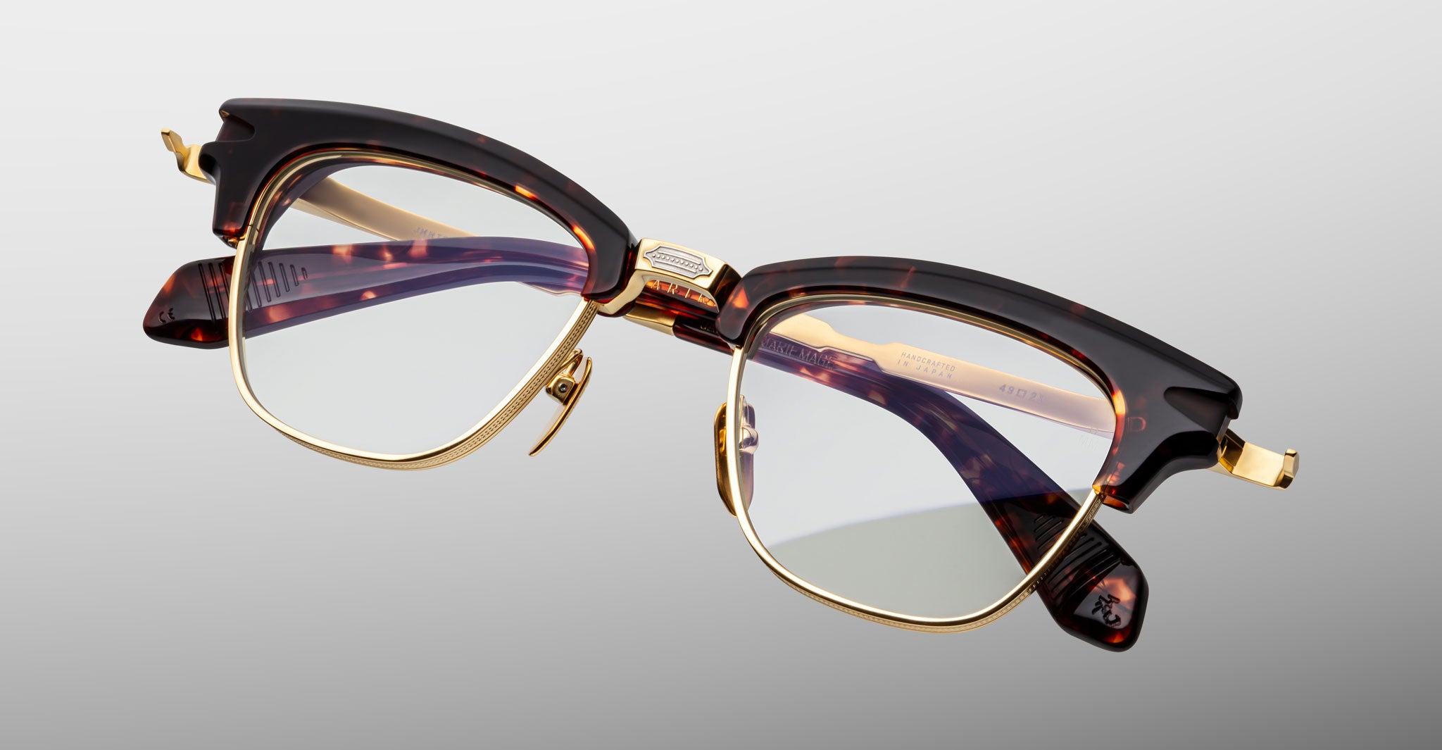 The Jacques Marie Mage Sartre glasses feature tortoiseshell-patterned clubmaster optical frames with gold accents and clear lenses, shown on a light gray background.